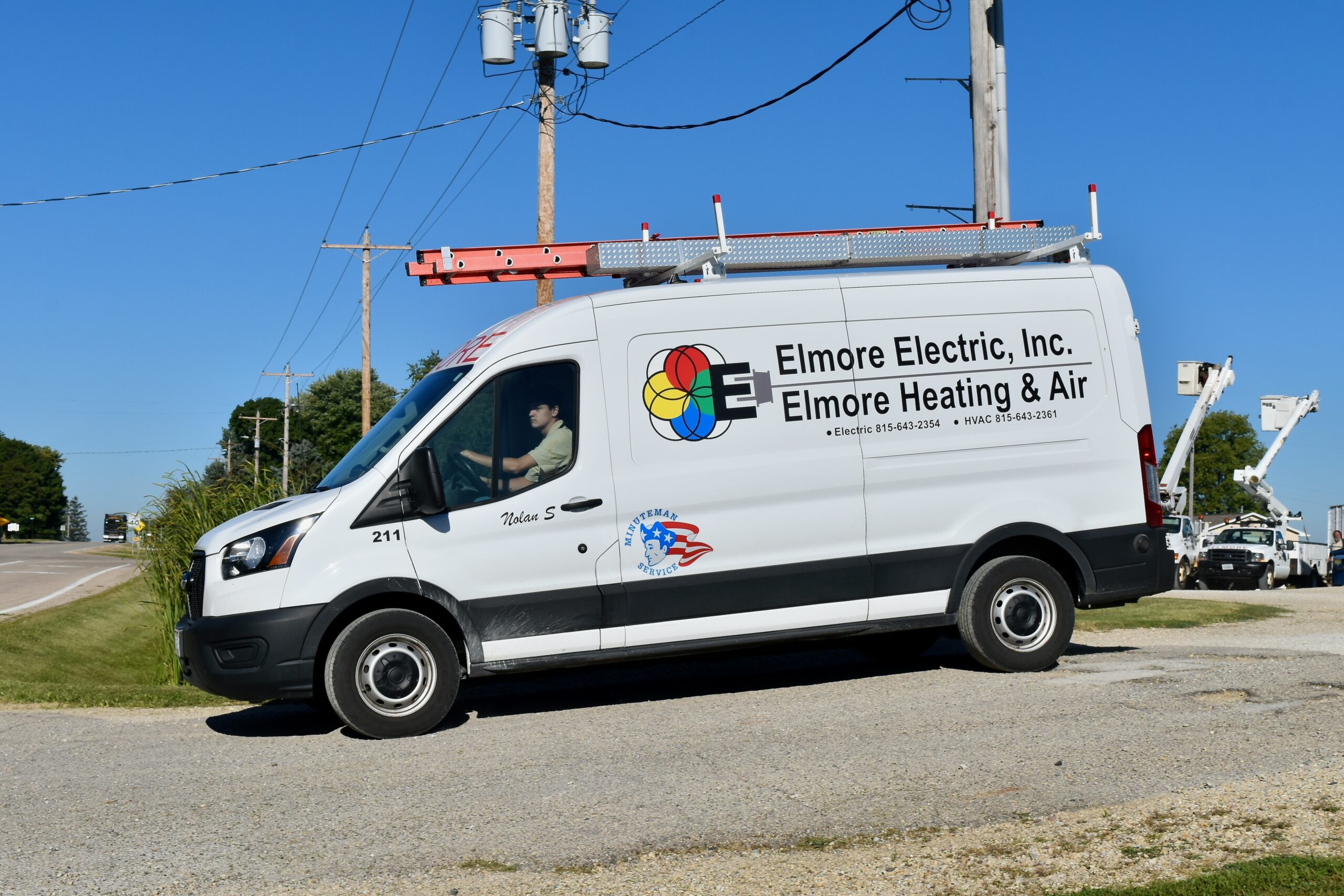 A white Elmore Electric, Inc. van with company logos and contact information is parked near a road with a clear blue sky in the background. The van is equipped with a ladder on top.